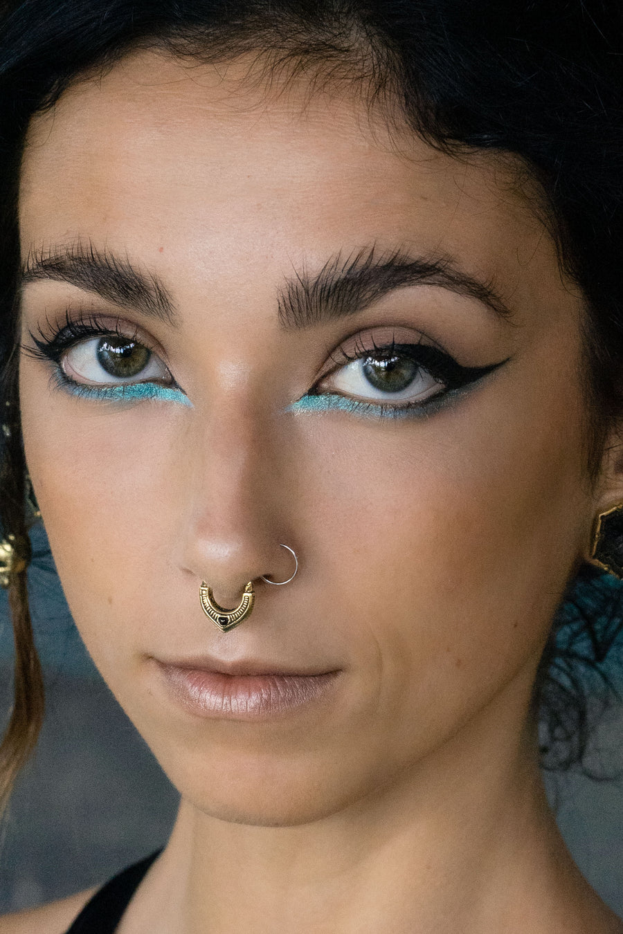 Close-up portrait of a woman with turquoise and black winged eyeliner, wearing a detailed gold septum ring with a circular design and a small gemstone. She gazes directly into the camera with a calm and confident expression.