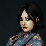 Side profile of a tattooed model wearing a biomechanical gold septum clicker and stretched ear plugs. Her piercing gaze and gothic-inspired attire enhance the post-apocalyptic fashion look.
