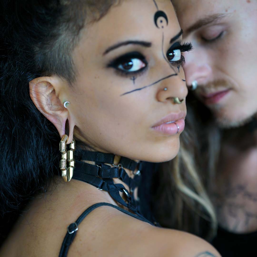 A woman with bold black face paint, large earrings, and piercings gazes intensely at the camera. She wears a black harness-style top and heavy gold TECHRE Futuristic Sci Fi Hexagon Keyhole Ear Weights from Kaya Movement. A man with facial hair and a slightly blurred appearance leans close to her in the background.