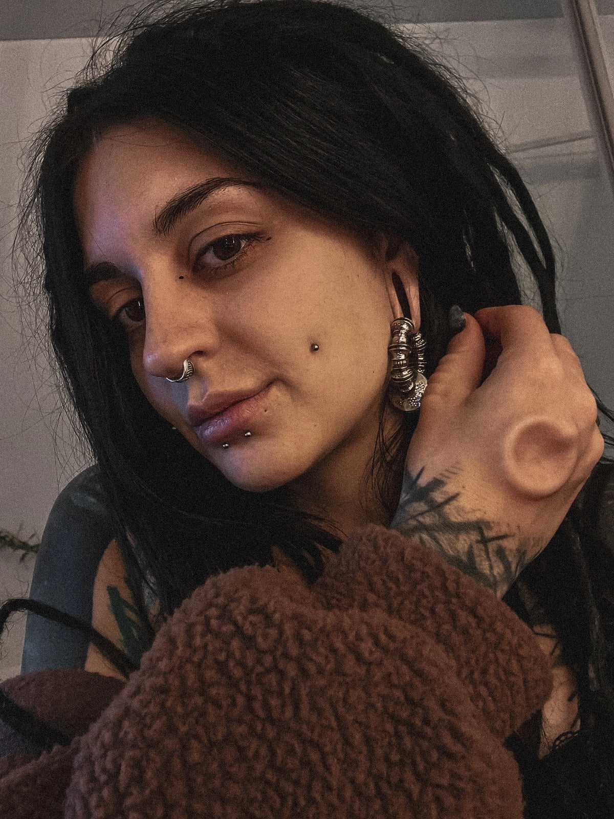 A person with long dark hair, visible septum, lip, and ear piercings adorned with Kaya Movement's TRAKARA Heavy Tribal Cyborg Clicker Ear Weights in Silver (2 gauge), is wearing a brown textured garment. They have a tattoo on their hand and wrist and are holding a lock of their hair while looking at the camera with a relaxed expression.