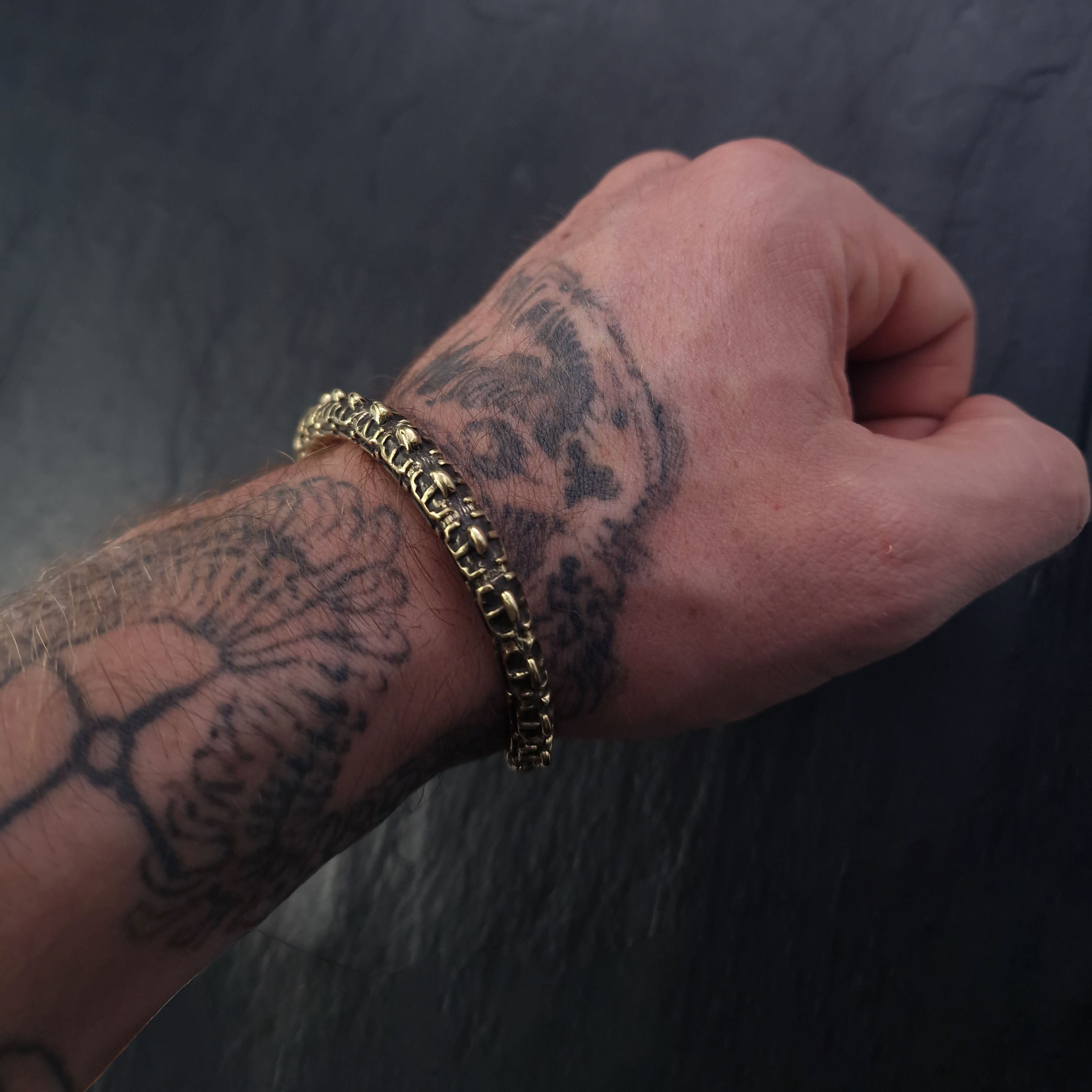 A close-up of a tattooed person's wrist wearing the SHIRO Men's Gold Cuff Bracelet by Kaya Movement. The tattoo features intricate designs, and the bracelet, with its reptile spine-inspired connected links, adds a contrasting metallic texture. The background is a dark, textured surface.