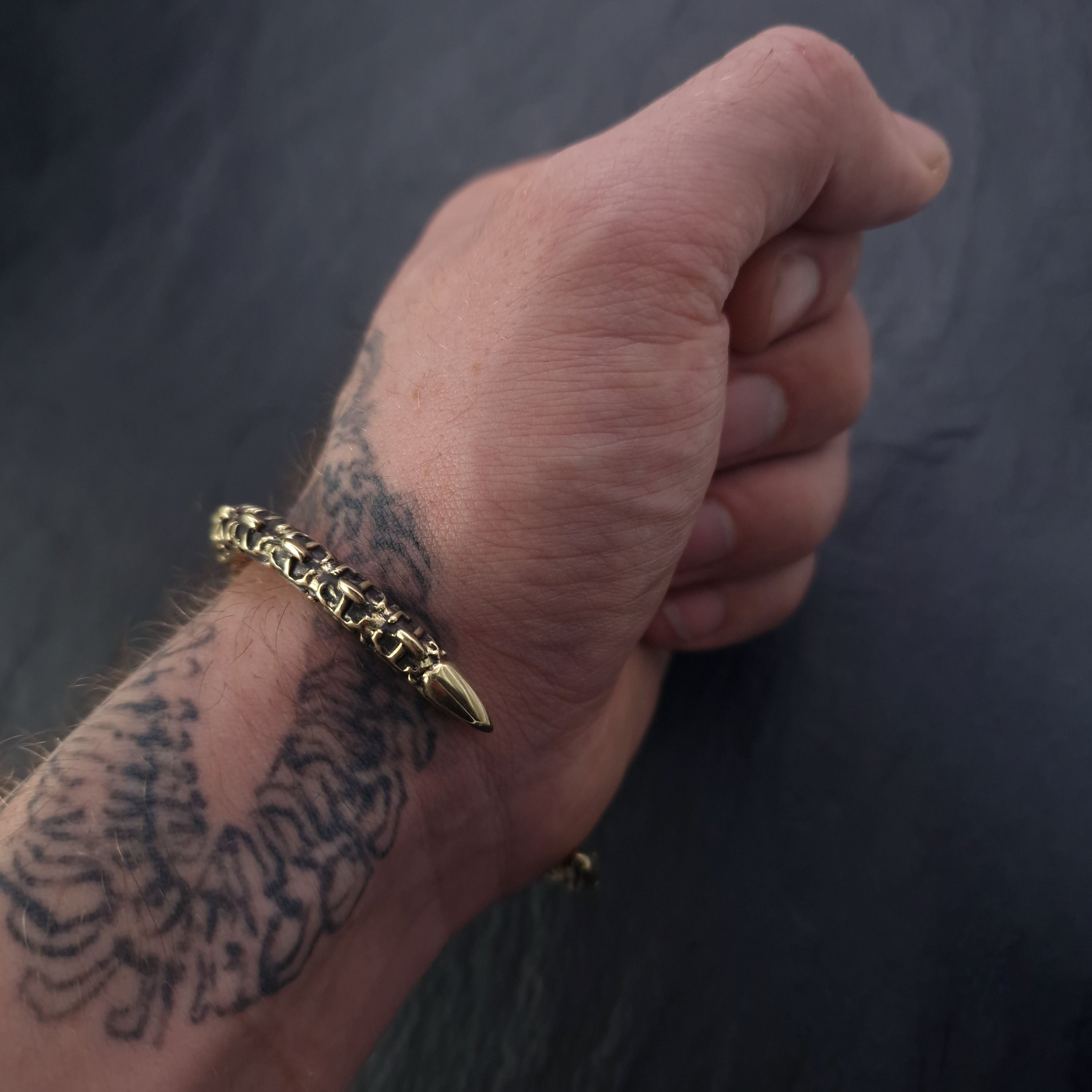 A close-up of a hand and wrist adorned with a detailed tattoo is showcased. The wrist flaunts the SHIRO Men's Gold Cuff Bracelet from Kaya Movement, featuring intricate designs, including a pointed charm that resembles a reptile spine design. The dark, blurred background accentuates the tattoo and bracelet.