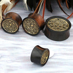 Close-up of four MAYA Round Saddle Plugs in Gold & Wood by Kaya Movement, ranging from 12mm to 25mm gauge, showcasing circular brass inlays with intricate geometric patterns. The double-flared saddle plugs are displayed on a light, textured surface, with feathers visible in the background.