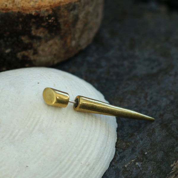 A close-up of the Kaya Movement PIKA Fake Talon Gauge Earrings in Gold with a 925 silver post (20 gauge) on a white shell, placed on a dark textured surface. The earring features a cylindrical end and cone-shaped body that tapers to a point, evoking a tribal boho style. Nearby, there is a blurred section of a brown, rocky object.