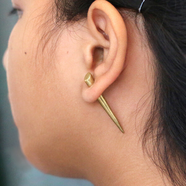 Close-up side view of a person's ear adorned with the PIKA Fake Talon Gauge Earrings in Gold by Kaya Movement, creating an illusion of a golden dagger piercing through the ear and capturing a tribal boho style. The pointed and elongated design with a slightly thicker base features subtle 925 silver wire accents on the 20 gauge post. The person's hair is pulled back, clearly revealing the unique earring.