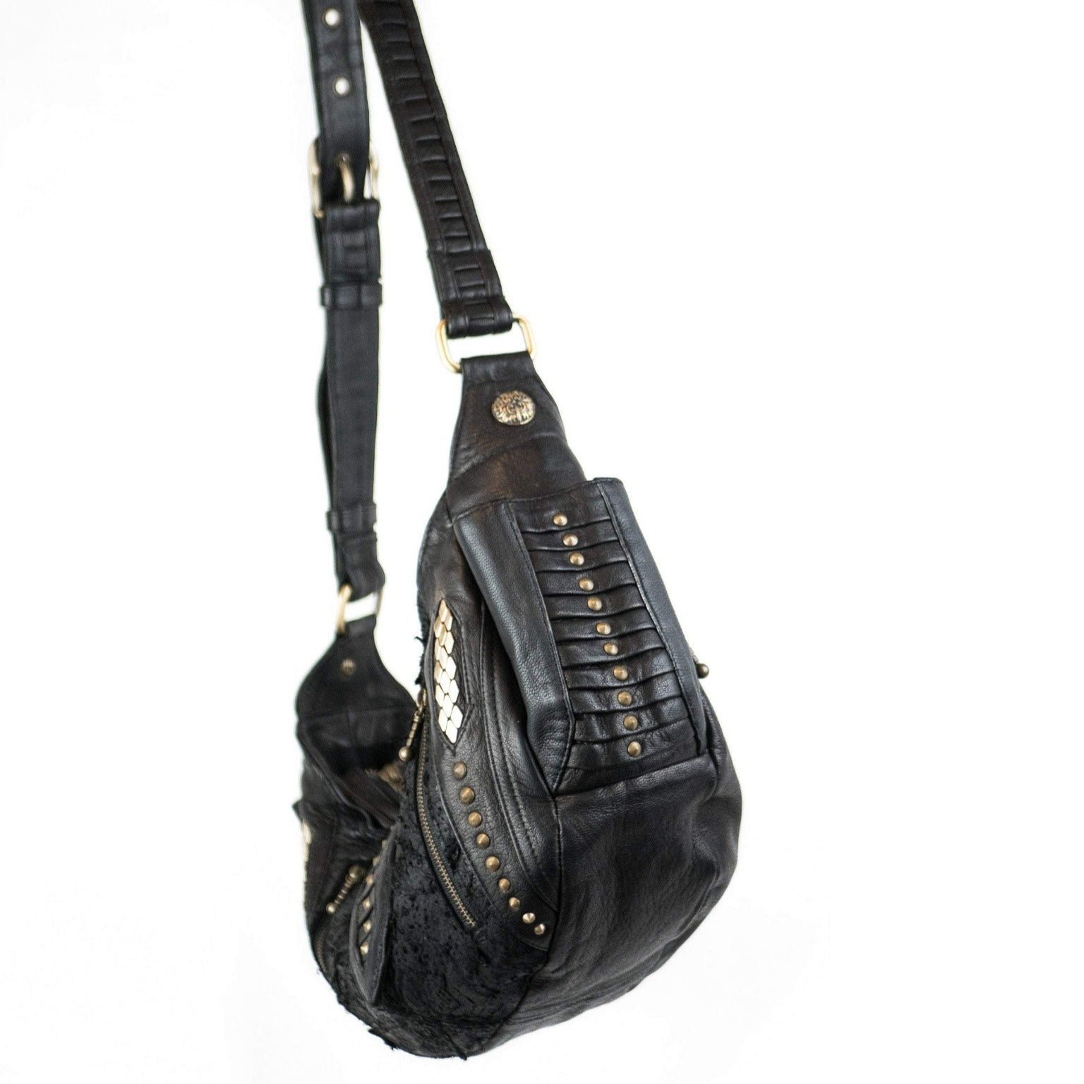 A black leather Nebula Unisex Leather Off Shoulder Cross Body HandBag from Kaya Movement, adorned with brass stud embellishments on the straps and sides, is hanging by its shoulder strap. This custom leather bag features decorative stitching and an asymmetrical design, giving it a unique, edgy appearance perfect for dystopian festivals.