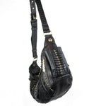 A black leather Nebula Unisex Leather Off Shoulder Cross Body HandBag from Kaya Movement, adorned with brass stud embellishments on the straps and sides, is hanging by its shoulder strap. This custom leather bag features decorative stitching and an asymmetrical design, giving it a unique, edgy appearance perfect for dystopian festivals.