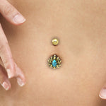 Close-up of a belly button adorned with the LIOSA Marquise Belly Piercing Ring in Gold & Turquoise by Kaya Movement, featuring a 14 gauge blue gem and an intricate, petal-like design. Two fingers gently touch the skin near the elegant belly jewelry.