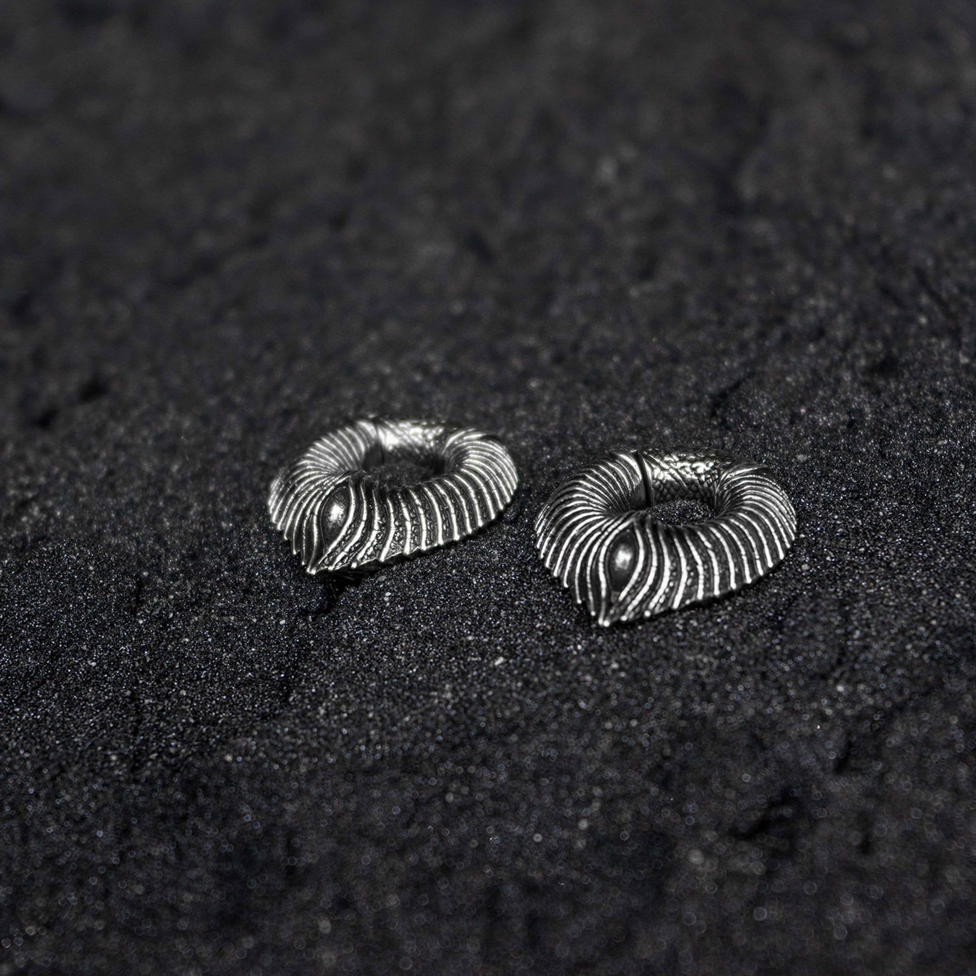 A pair of MARVDASHT Medium Cyborg Clicker Ear Weights in Silver Brass from Kaya Movement lies on a dark, grainy surface. These earrings, perfect for stretched ears at a 3/8" gauge, feature an intricately designed ribbed pattern that gives them a textured, striped appearance.