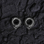 On a textured black surface lie two intricately designed, metallic, ring-shaped objects. Resembling curled, ornate serpents, these MARVDASHT Medium Cyborg Clicker Ear Weights from Kaya Movement feature detailed patterns with pointed ends touching the ground. Made of 925 silver and measuring 3/8" gauge, the dark and granular backdrop enhances their dystopian body jewelry aesthetic.