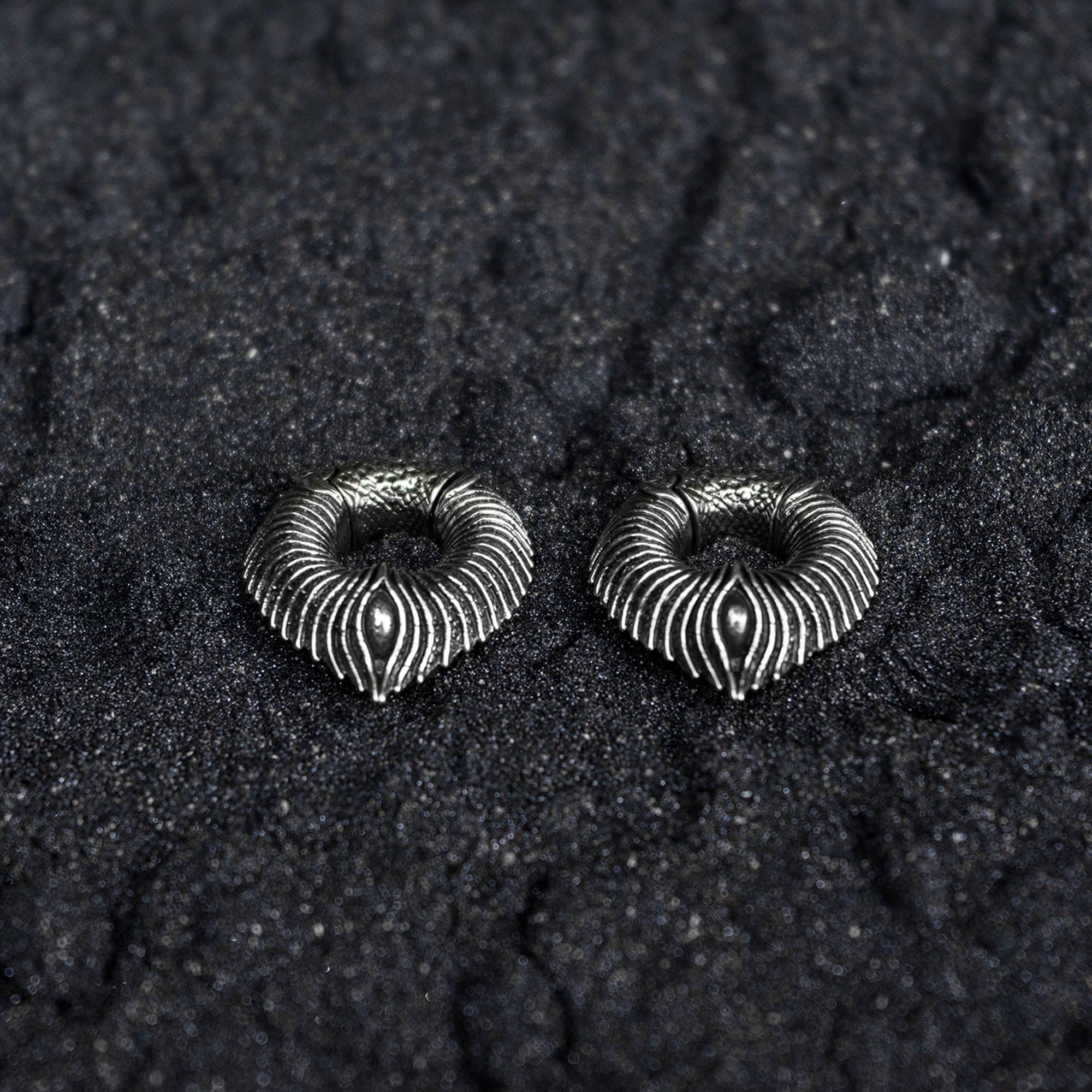 Two intricately designed silver-colored MARVDASHT Medium Cyborg Clicker Ear Weights in Silver Brass from Kaya Movement rest on a textured, dark black surface. Featuring a symmetrical, fan-like pattern with fine engraved lines radiating outward, these 3/8" gauge pieces evoke the sophisticated elegance of dystopian body jewelry.