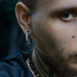 Close-up of the right side of a man's face, showcasing the MARVDASHT Small Biomechanical Clicker Ear Weights in 925 Silver (2 gauge) by Kaya Movement, beautifully nestled in his ear and giving major sci-fi vibes. He sports tattoos near his ear and on his neck, paired with a beaded necklace around his neck. The dark, out-of-focus background adds to the futuristic feel.