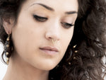 Close-up of a person with curly brown hair looking down, wearing the KAMO Biomech Teardrop Septum Ring in Gold by Kaya Movement and large hoop earrings adorned with beads and spikes. The expression is thoughtful, and the background is neutral.