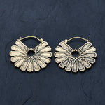 A pair of MOSAIC Ethnic Mandala Disc Hoop Earrings in Gold by Kaya Movement lay on a dark slate surface. Each 16-gauge earring features a radially symmetrical, sunburst design with textured, petal-like segments radiating from a central hole, exuding the charm of ethnic hoop earrings and creating a striking and elegant appearance.