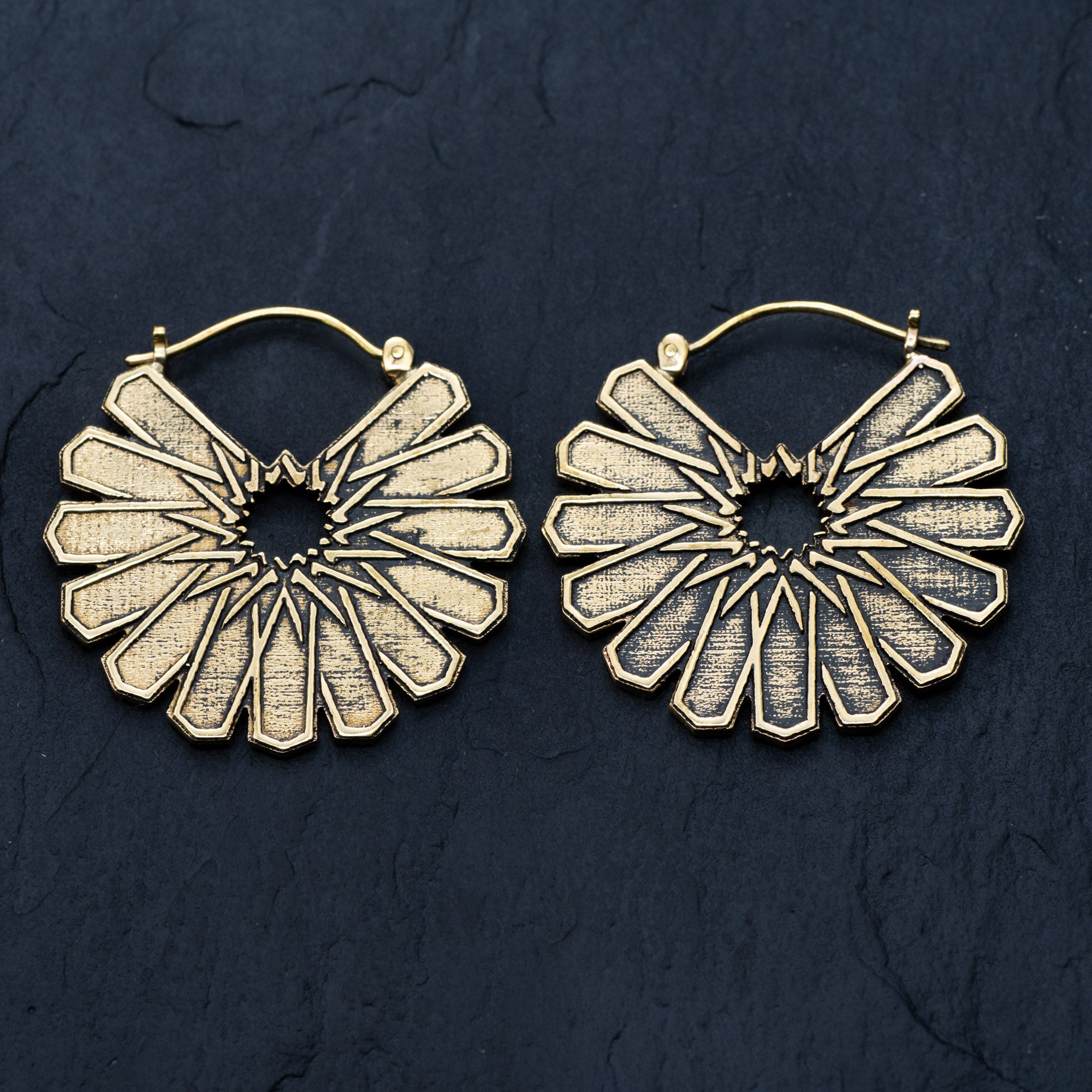 Presenting the MOSAIC Ethnic Mandala Disc Hoop Earrings in Gold by Kaya Movement. These 16-gauge boho statement earrings boast a beautifully intricate radial pattern, reminiscent of a starburst or sunburst, placed against a dark textured background. Featuring a latch back closure and detailed cut-out designs, they exude an elegant and vintage charm.