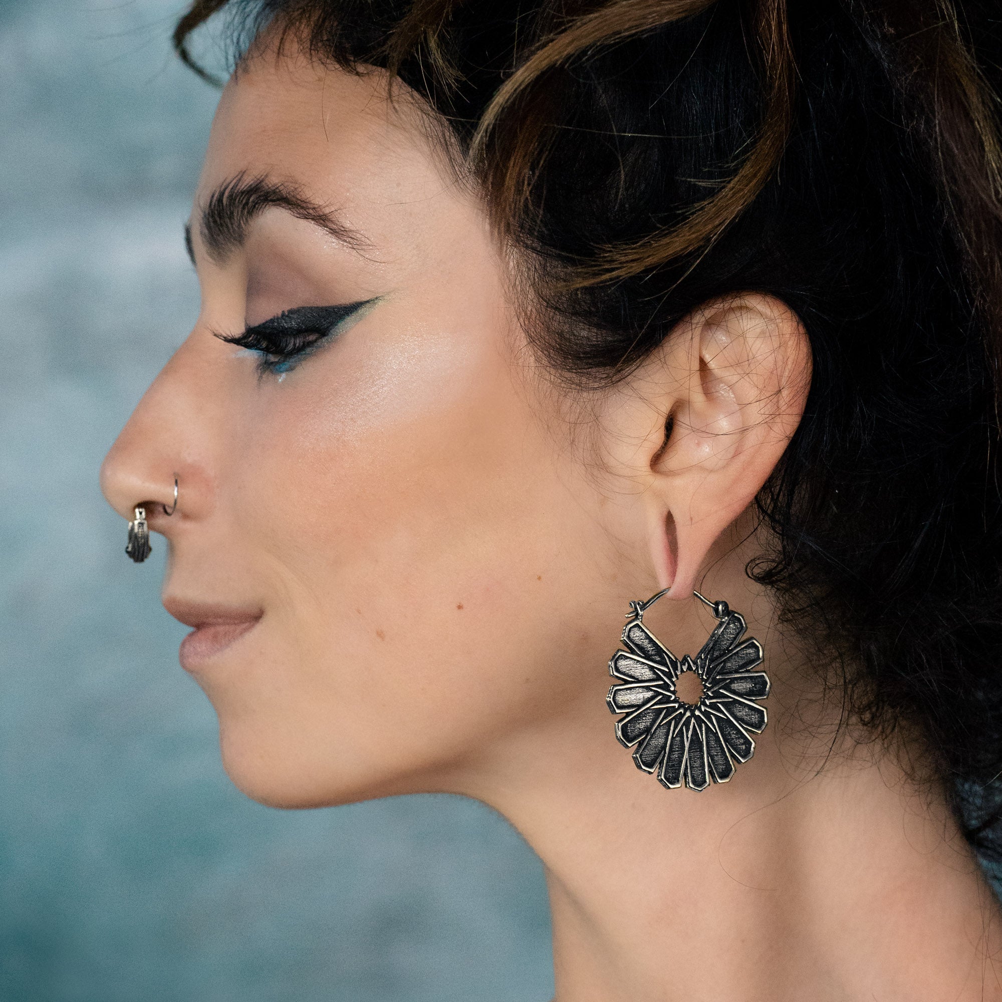 A person with winged eyeliner and a nose piercing is shown in profile, showcasing Kaya Movement's MOSAIC Ethnic Mandala Disc Hoop Earrings in Silver, crafted at 16 gauge. The blurred background with a blue and grey gradient highlights the intricate floral design of the metallic hoops.