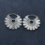 A pair of MOSAIC Ethnic Mandala Disc Hoop Earrings in Silver (16 gauge) by Kaya Movement is laid on a dark textured surface. Each earring features radiating triangular segments forming a circular pattern with a cut-out central area, echoing the essence of boho statement earrings.