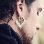 Close-up of a person's ear adorned with a handcrafted CROW SKULL Gothic Silver Ring with Gold Charm by Kaya Movement, made from 316L surgical steel. The geometric, bent metal shape is accented by a gold brass raven skull pendant. The person has long, dark hair tied back and is dressed in a dark garment.