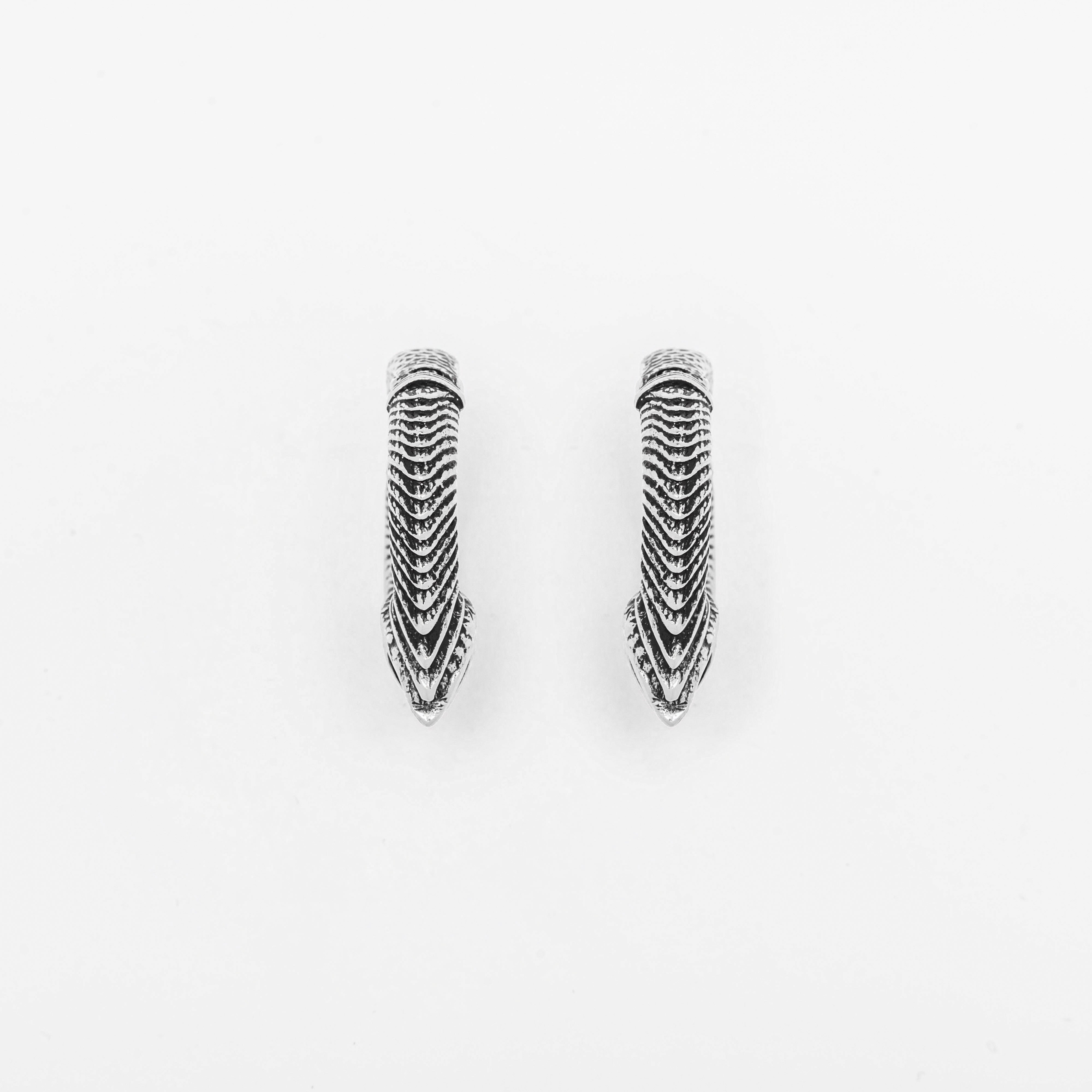 The MARVDASHT Small Biomechanical Clicker Ear Weights in Silver Brass by Kaya Movement are displayed on a white background. These 2-gauge ear weights have an elongated, pointed shape with intricate, layered detailing that creates a textured, feather-like appearance—reminiscent of elegant post-apocalyptic body jewelry.
