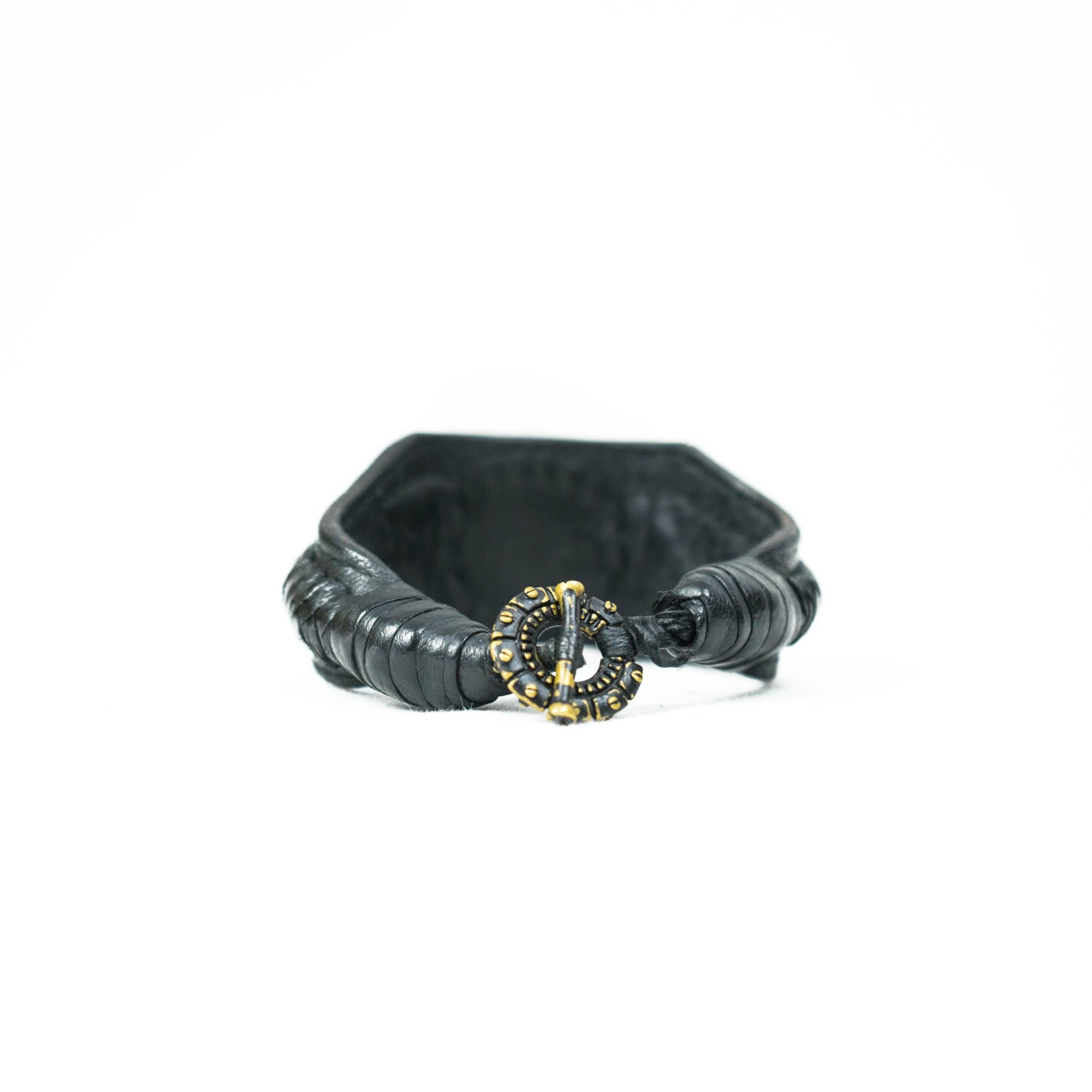 The ANOM Men's Black Cuff Leather Bracelet by Kaya Movement showcases textured, braided leather with an ornate golden clasp adorned with genuine Amethyst gemstone elements, secured at the front. Positioned against a plain white background, this piece embodies elegance and style.