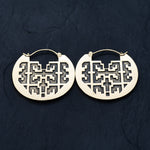 The CONIBOS Tribal Shipibo Hoop Disc Earrings in Gold by Kaya Movement, a pair of 16-gauge earrings with intricate and symmetrical cut-out patterns, are laid on a dark, textured surface. Their design features geometric shapes and lines creating a visually appealing and traditional look that embodies the essence of tribal earrings.