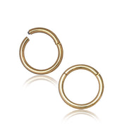 Two small, polished AMOI Clicker Nose Rings in gold, crafted from surgical steel and gold-coated by Kaya Movement, are displayed on a white background. The multi-use piercing rings have simple, smooth surfaces with a hinge mechanism for easy opening and closing. One ring is slightly open, showing the clasp. A subtle shadow is visible beneath them.