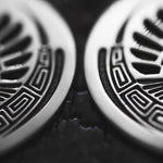 Close-up image of two CROP CIRCLE Oversized Tribal Disc Hoop Earrings in Silver by Kaya Movement, showcasing intricate black and white geometric patterns on a dark surface. The design features a mix of straight lines and curves, creating an abstract, symmetrical pattern that appears metallic.