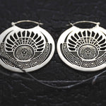 The CROP CIRCLE Oversized Tribal Disc Hoop Earrings in Silver from Kaya Movement are intricately designed, 16-gauge circular earrings featuring a tribal sun motif against a textured black background. These boho body jewelry pieces showcase geometric patterns and are symmetrically arranged.