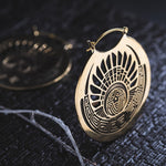 Close-up of two intricate CROP CIRCLE Oversized Tribal Disc Earrings in Gold by Kaya Movement, featuring detailed geometric and tribal-style patterns. The earrings rest on a textured dark surface, with one slightly out of focus in the background. A small, delicate branch is also visible.