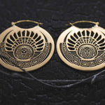 A pair of CROP CIRCLE Oversized Tribal Disc Earrings in Gold by Kaya Movement, boasting intricate cut-out designs featuring an abstract sun motif at the center and geometric patterns around the edges. These 16-gauge, tribal statement earrings are set against a textured, dark background.