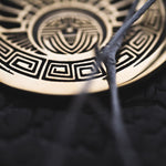 Close-up image of a circular, gold-colored metal ornament featuring CROP CIRCLE Oversized Tribal Disc Earrings in Gold by Kaya Movement. The intricate geometric patterns include a labyrinth-like design and a stylized face. Two dry twigs partially cover the 16-gauge earrings, evoking the style of oversized tribal statement pieces against the dark background.