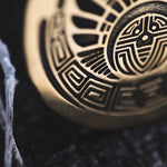Close-up of the intricately designed CROP CIRCLE Oversized Tribal Disc Earrings in Gold by Kaya Movement, resting on a dark textured surface. The earrings feature geometric patterns and abstract shapes resembling a face or mask, capturing the fine details and craftsmanship of this 16-gauge piece.