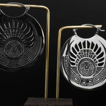 The CROP CIRCLE Oversized Tribal Disc Hoop Earrings in Silver by Kaya Movement are a pair of round, metallic gauge earrings featuring intricate geometric and tribal designs with a central face element. One earring appears in silver, while the other has a darker, possibly oxidized finish. Displayed on a gold and black stand against a black background, this boho body jewelry makes an enchanting statement.