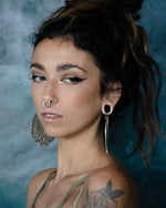 A person with dark, wavy hair styled in a loose updo is adorned with Kaya Movement's Circuit Oversized Visionary Disc Hoop Earrings in Silver (16 gauge) and a septum ring. Their bold eyeliner makeup complements their boho statement look. Sporting a tattoo on their upper arm, they complete the ensemble with a strappy top against a teal background.