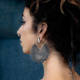 Profile view of a person with dark hair tied up in a messy bun, wearing the Circuit Oversized Visionary Disc Hoop Earrings in Silver (16 gauge) by Kaya Movement and a small nose ring. The person also has a tattoo on their back and is dressed in a light-colored tank top. The boho statement look is set against a blurred, dark blue background.