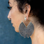 A person with dark hair wears the Kaya Movement’s Circuit Oversized Visionary Disc Hoop Earrings in Silver, 16 gauge, which are intricately designed and shaped like a circular labyrinth, making a stunning boho statement. They also have a nose ring. The background is a textured blue surface.