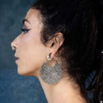 A woman with dark hair styled in a loose ponytail is shown in profile against a dark, textured background. She is wearing Kaya Movement's PANCHA Oversized Tribal Shipibo Disc Earrings in Silver, which feature elaborate and intricate patterns, along with a nose ring. Her eye makeup is highlighted by bold, winged eyeliner.