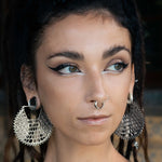 A person with dark braided hair wears the bold Kaya Movement ILLUSION Oversized Cyborg Sci Fi Disc Earrings in Silver, complete with intricate patterns. Complementing the earrings are a septum ring and multiple ear piercings. Their makeup features striking eyeliner extending outward and white accents below the eyes, creating a distinct sci-fi aesthetic as they gaze into the distance.