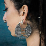 A person with long, dark hair is depicted in profile, showcasing the ILLUSION Oversized Cyborg Sci Fi Disc Earrings in Silver by Kaya Movement. These 14-gauge earrings feature an intricate honeycomb pattern with spiked edges, giving off a strong sci-fi aesthetic. The individual also wears a nose ring and several ear piercings. The background is a subdued teal hue.