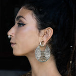 A person with long dark hair tied back is shown in profile view. They wear a nose ring and large, intricate ILLUSION Oversized Cyborg Sci Fi Disc Earrings in Silver from Kaya Movement, exuding strong sci-fi vibes. The makeup on their eyes is bold, featuring sharp black eyeliner. The background is dark and out of focus.