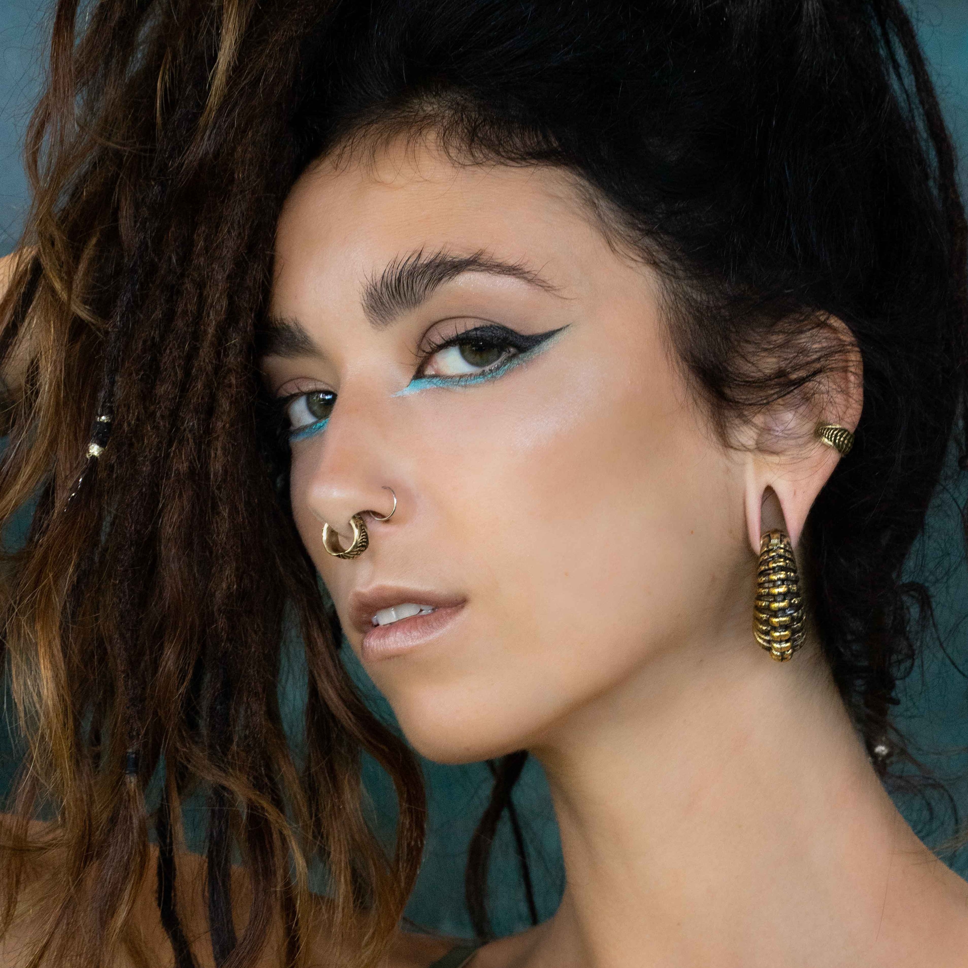 A person with dark hair styled in dreadlocks stares into the camera with a serious expression. They have bold blue and black eyeliner, unisex nose jewelry in the septum from Kaya Movement's DARMA Fake Septum Ring or Ear Cuff in Gold, and large gold earrings shaped like pinecones. Their background is a soft, blue-green gradient.
