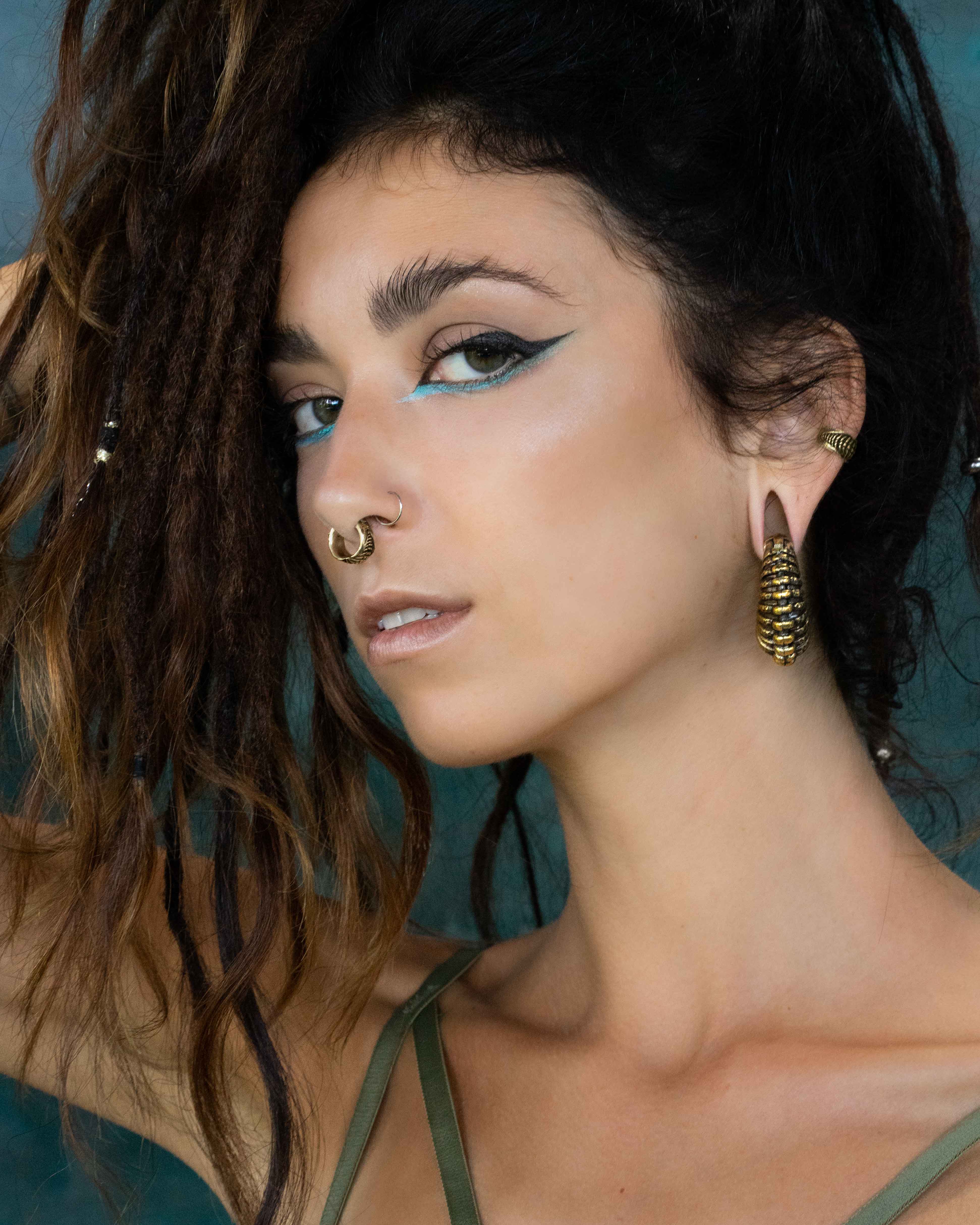 A woman with long, dark dreadlocks adorned with beads poses confidently with one hand in her hair. She has bold eyeliner with a blue accent, a septum piercing, and wears the TRILOBITE Large Biomechanical Clicker Ear Weights in Gold Brass by Kaya Movement at 0 gauge. She is dressed in a green top.