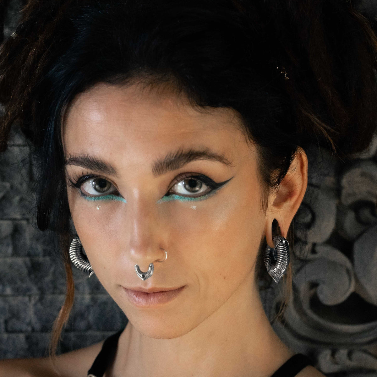 A portrait capturing an individual with dark hair, bold black eyeliner, and striking blue eye shadow. They sport a nose ring and prominently wear Kaya Movement's MARVDASHT Medium Cyborg Clicker Ear Weights in Silver Brass (3/8" gauge) in their stretched ears. The backdrop is a textured, stone-like wall. The person gazes directly at the camera with a neutral expression.