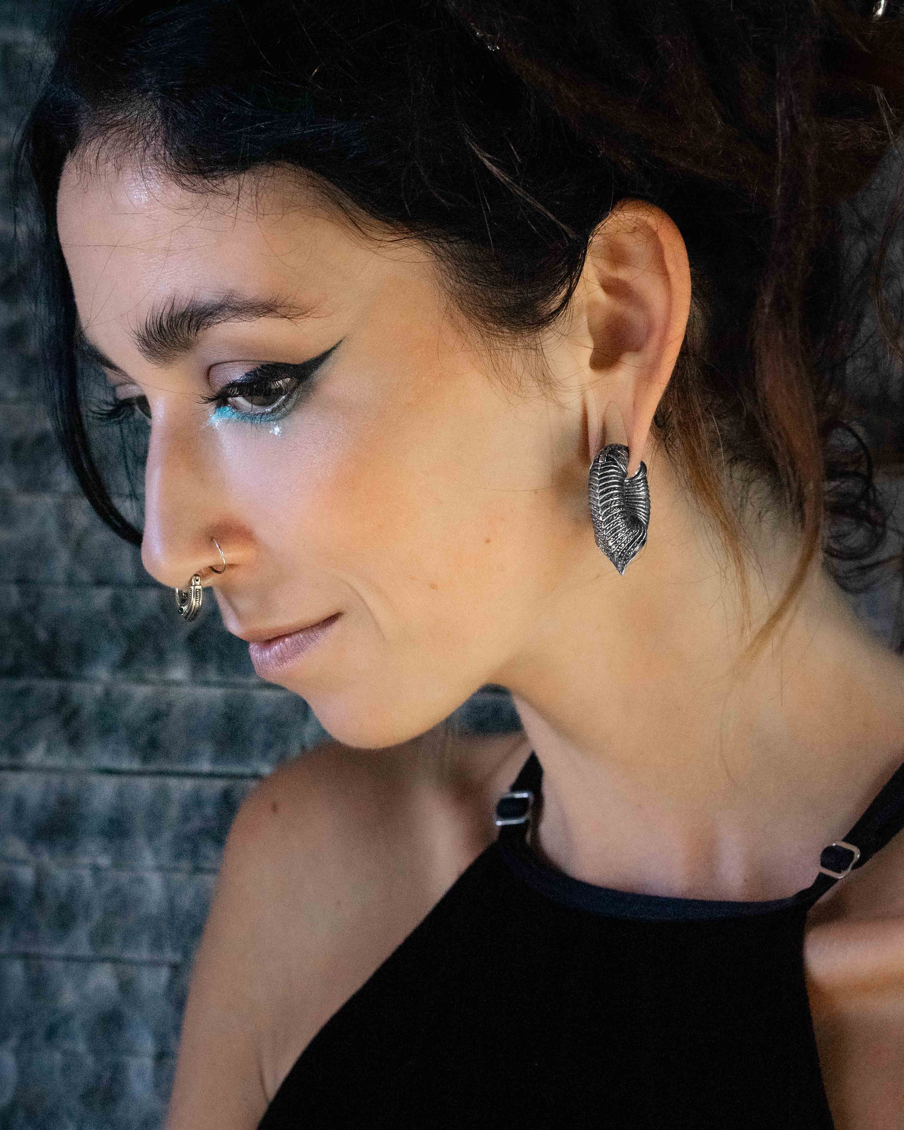 A person with dark hair and dramatic winged eyeliner is looking down. They are wearing a black top with thin straps and have a small nose ring. Their jewelry includes the MARVDASHT Medium Cyborg Clicker Ear Weights in Silver Brass (3/8" gauge) by Kaya Movement, which are large, textured, and heart-shaped, reminiscent of dystopian body jewelry. The background is a textured, dark surface.