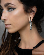A person with long dark hair and an intricate nose ring is wearing bold silver hoop earrings and dramatic aqua blue eyeliner. They are dressed in a black top with metal ring accents and post-apocalyptic body jewelry, including the unique Kaya Movement TECHRE Futuristic Hexagon Keyhole Ear Weights in Silver (0 gauge). The neutral background highlights their accessories and makeup.