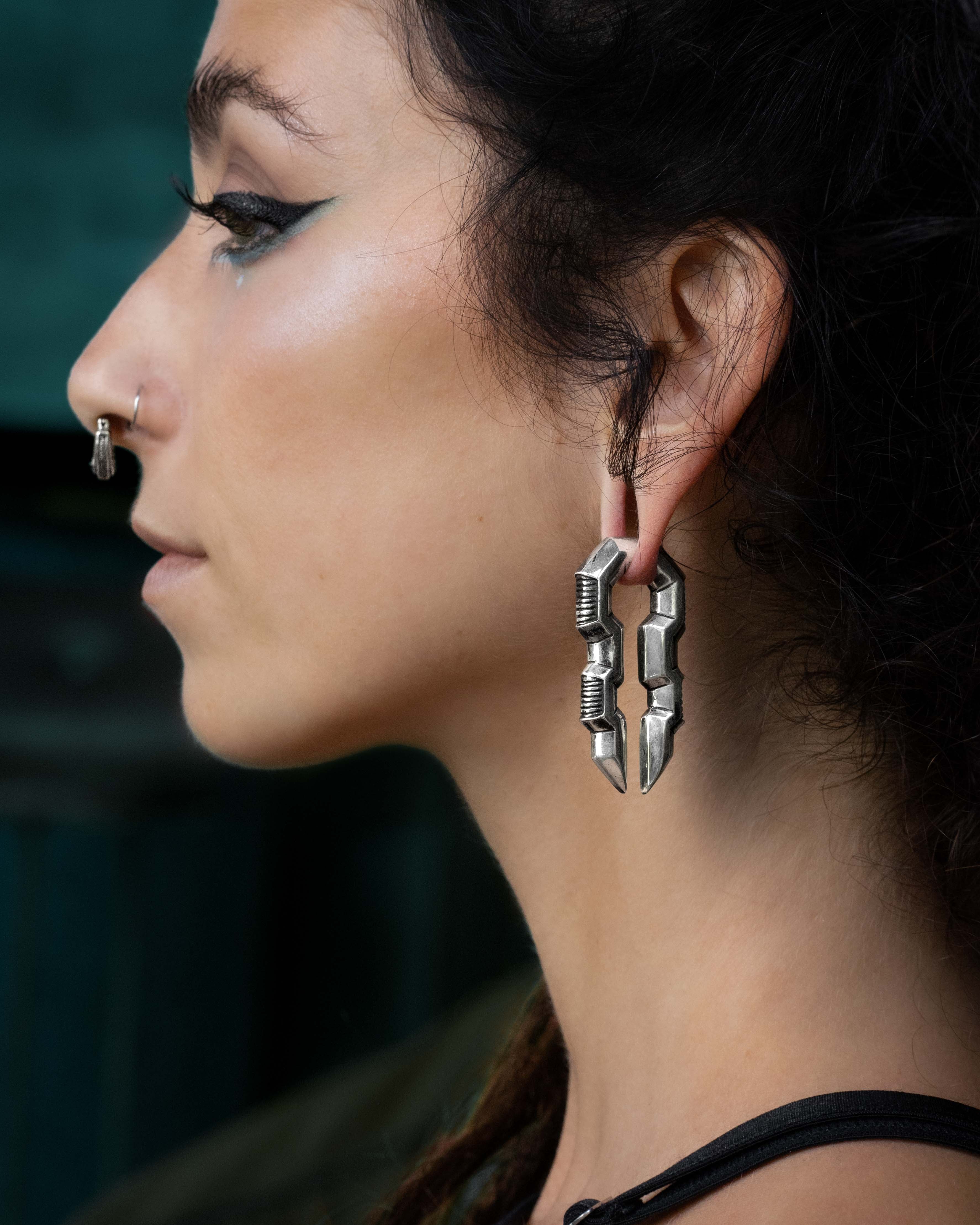 A person with dark hair and winged eyeliner is shown in profile. They are wearing Kaya Movement's TECHRE Futuristic Hexagon Keyhole Ear Weights in Silver (0 gauge), which resemble interlocking segments of post-apocalyptic body jewelry, along with a silver nose ring. The background is out of focus, emphasizing the jewelry and their side profile.