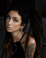 A person with long, dark dreadlocks tied in two ponytails is wearing a sleeveless black top. They have a septum piercing and are adorned with Kaya Movement’s TECHRE Futuristic Hexagon Keyhole Ear Weights in Silver (0 gauge), along with blue eye makeup. A tattoo of a rose is visible on their upper arm. The background is dark and out of focus.