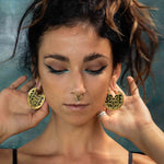 A person with dark makeup and winged eyeliner holds up the large CONIBOS Tribal Shipibo Hoop Disc Earrings in Gold from Kaya Movement against their ears. They have a nose ring, and their hair is loosely tied up, with some loose strands framing their face. The boho body jewelry stands out against the muted, textured background.