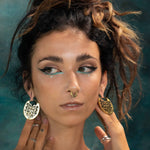 A person with dark hair styled in messy updos, adorned with dramatic winged eyeliner and a nose ring. They are wearing the CONIBOS Tribal Shipibo Hoop Disc Earrings in Gold by Kaya Movement, along with multiple rings on their fingers, posing with their hands near their face against a blurred turquoise background.