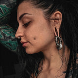 A person with dark hair and various piercings, including a unisex Kaya Movement Darma Biomechanical Clicker Hoop Septum Nose Ring in Silver (16 gauge), ear gauges, and a dermal piercing on the cheek, poses with one arm raised, showcasing their profile. They have tattoos visible on their arm. The background is dark and out of focus.